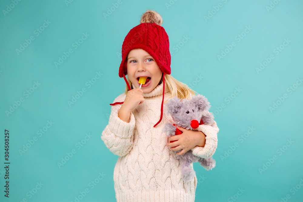 little blonde girl in a red knitted hat and sweater holding a soft toy mouse and a yellow Lollipop. isolate on blue background, space for text - obrazy, fototapety, plakaty 