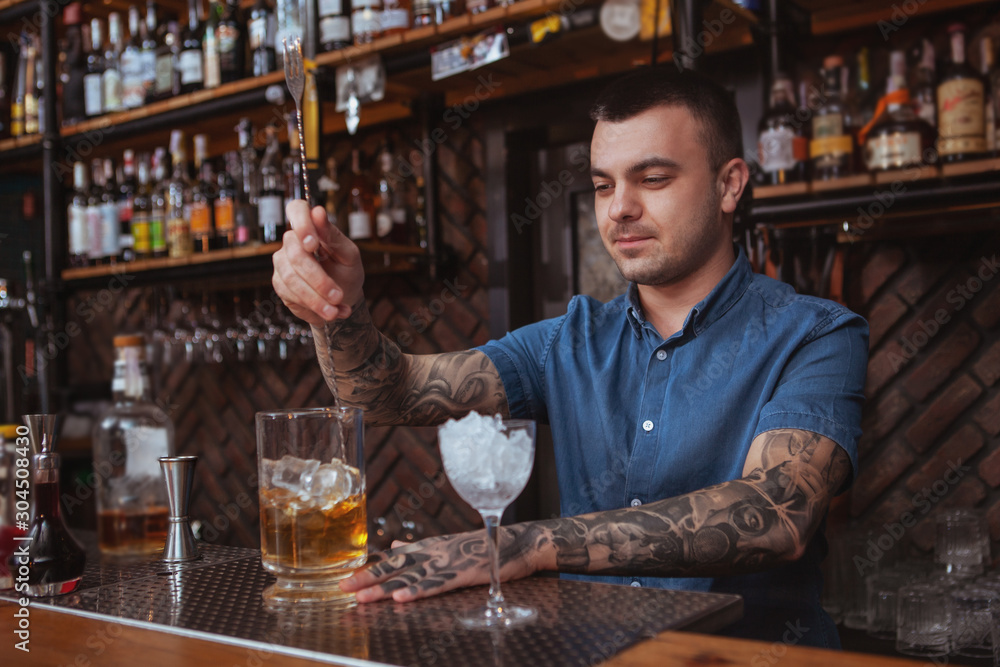 Handsome tattooed bartender smiling, mixing alcohol, preparing cocktails