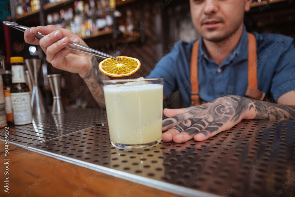 Cropped close up of a tattooed bartender preparing delicious cocktail, garnishing the drink with orange slice Cropped close up of a tattooed bartender preparing delicious cocktail, garnishing the drink with orange slice