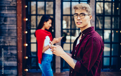 Pondering chinese hipster guy in eyeglasses looking away while messaging on smartphone and using 4G internet for communication with friend which standing on blurred background with cellular in hand