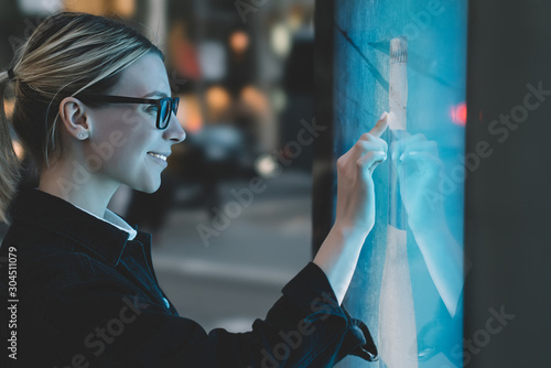 Smiling female standing at big display with advanced digital technology. Young woman touching with finger sensitive screen of interactive kiosk for find information while standing on street in evening photo