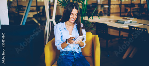 Cheerful hipster girl watching comedy show on website using digital tablet and free wireless connection, smiling woman browsing internet on modern touch pad while sitting in comfortable chair indoors photo