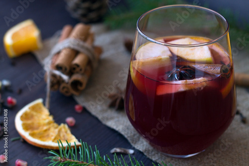 Hot mulled wine in a glass on a wooden background with apple and orange slices and spices with cinnamon. winter and autumn hot drinks. christmas still life with candle and decorations