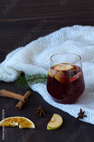 winter still life. glass of red mulled wine with orange, apple and cinnamon, anise star and warm white knitted plaid on a dark wooden background with green pine branch. with copy space