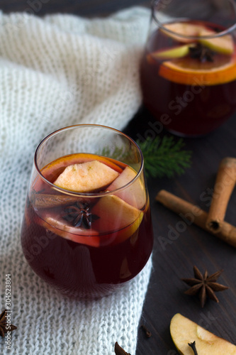 glass of red mulled wine with orange, apple and cinnamon, anise star and warm white knitted plaid on a dark wooden background with green pine branch. winter still life. close up