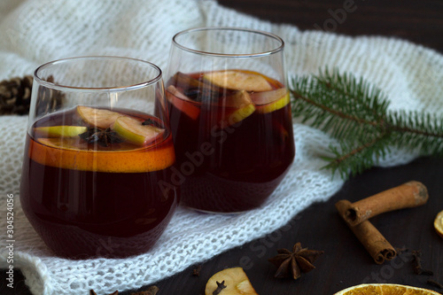 winter collection. tasty mulled wine in a two glasses with orange and cinnamon, anise star and warm white plaid on a dark wooden background with green pine branch