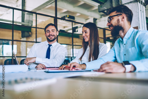 Portrait of cheerful male employee looking at camera while colleagues watching motivation webinar about partnership using wireless connection on laptop device, group of business people reading news