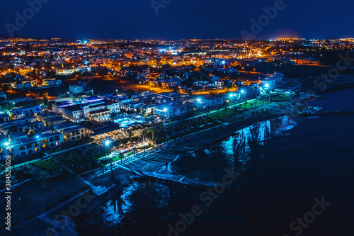 Aerial panoramic view of Paphos, Cyprus seaside from above at night from drone. Beautiful evening mediterranean seascape with illuminated buildings and reflections of lights in sea water. © DedMityay