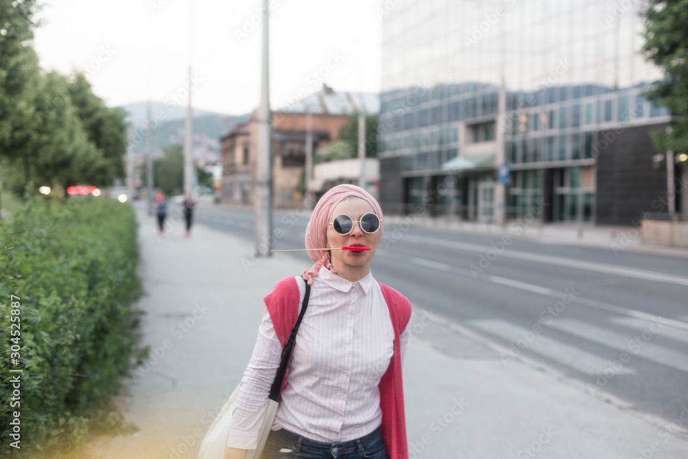 Fototapeta premium romantic portrait of young European Muslim woman with hijab have fun at beautiful sunny day. Image of pretty woman walking with red heart shaped lollipop. She is happy and relaxed.