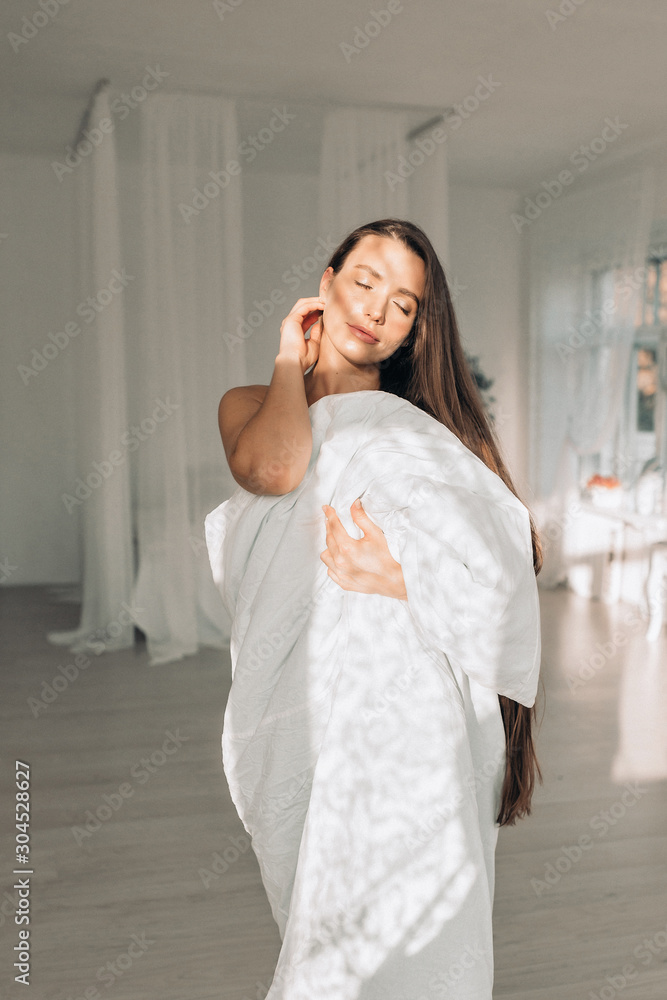 A beautiful long haired woman standing in white blanket in light room A beautiful long haired woman standing in white blanket in light room