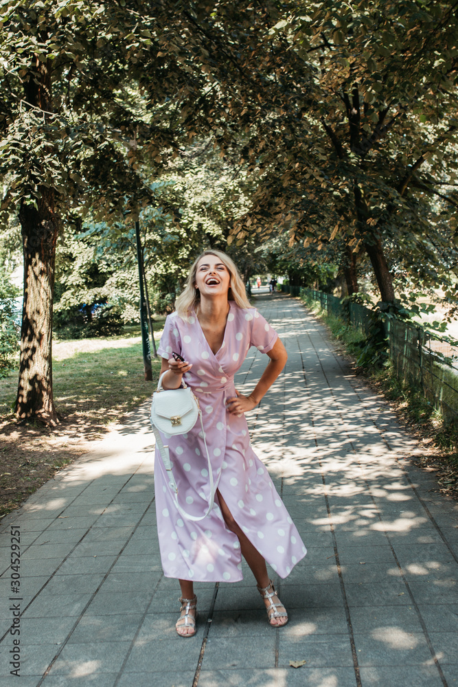 Beautiful stylish blonde woman in polka dotted midi dress in city summer park. She is happy and enjoying walk in sunny day. She is wearing small white bag and black sunglasses. Beautiful stylish blonde woman in polka dotted midi dress in city summer park. She is happy and enjoying walk in sunny day. She is wearing small white bag and black sunglasses.