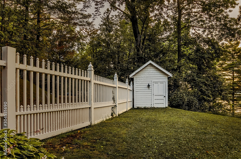 A white garden shed sits on a lawn with a white picket fence leading away from it on the edge of Lake Missawappi at sunrise A white garden shed sits on a lawn with a white picket fence leading away from it on the edge of Lake Missawappi at sunrise