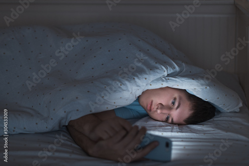Sleepy depressed teenager surfing in the internet on his mobile phone lying on bed in the dark. Web addiction, smartphone dependency, internet trap. Insomnia, nomophobia, sleep disorder concept.