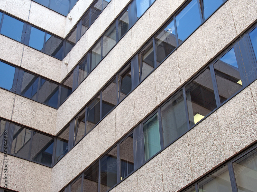 angled corner of a large concrete office building with other modern buildings and blue sky reflected in rows of windows angled corner of a large concrete office building with other modern buildings and blue sky reflected in rows of windows