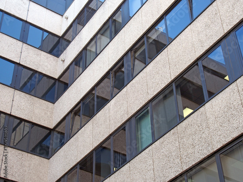 angled corner of a large concrete office building with other modern buildings and blue sky reflected in rows of windows