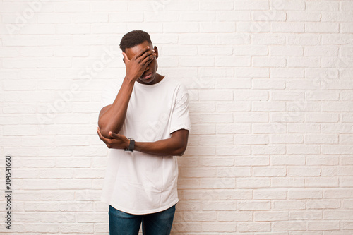 young african american black man looking stressed, ashamed or upset, with a headache, covering face with hand against brick wall photo