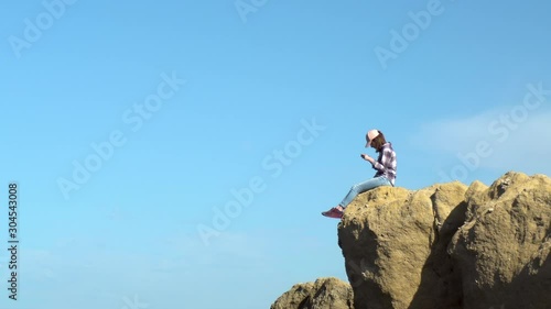 A young woman is sitting on a rock with a telephone. A woman in a cap dangled her legs over a cliff. Communication in the messenger on a smartphone photo