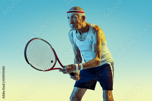 Senior man wearing sportwear playing tennis on gradient background, neon light. Caucasian male model in great shape stays active, sportive. Concept of sport, activity, movement, wellbeing, confidence. © master1305
