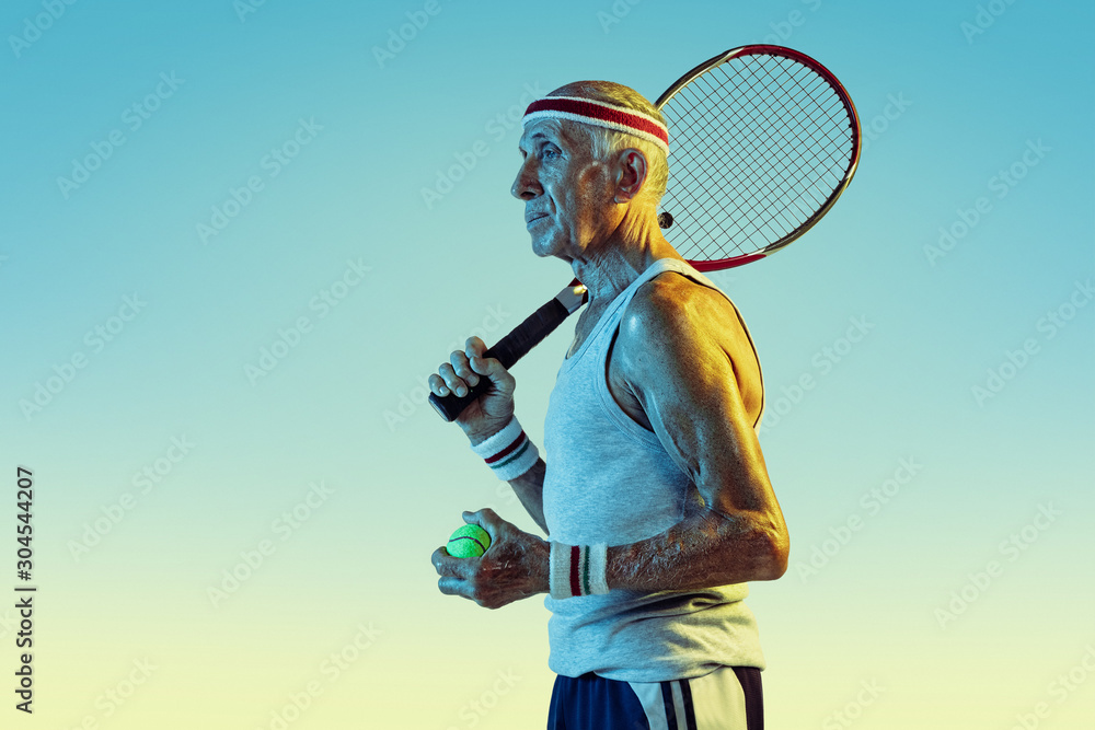 Senior man wearing sportwear playing tennis on gradient background, neon light. Caucasian male model in great shape stays active, sportive. Concept of sport, activity, movement, wellbeing, confidence.