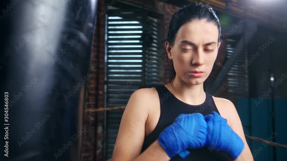 Aggressive female kickboxer with drops of sweat on face in blue wrapped hands posing at dark gym Aggressive female kickboxer with drops of sweat on face in blue wrapped hands posing at dark gym