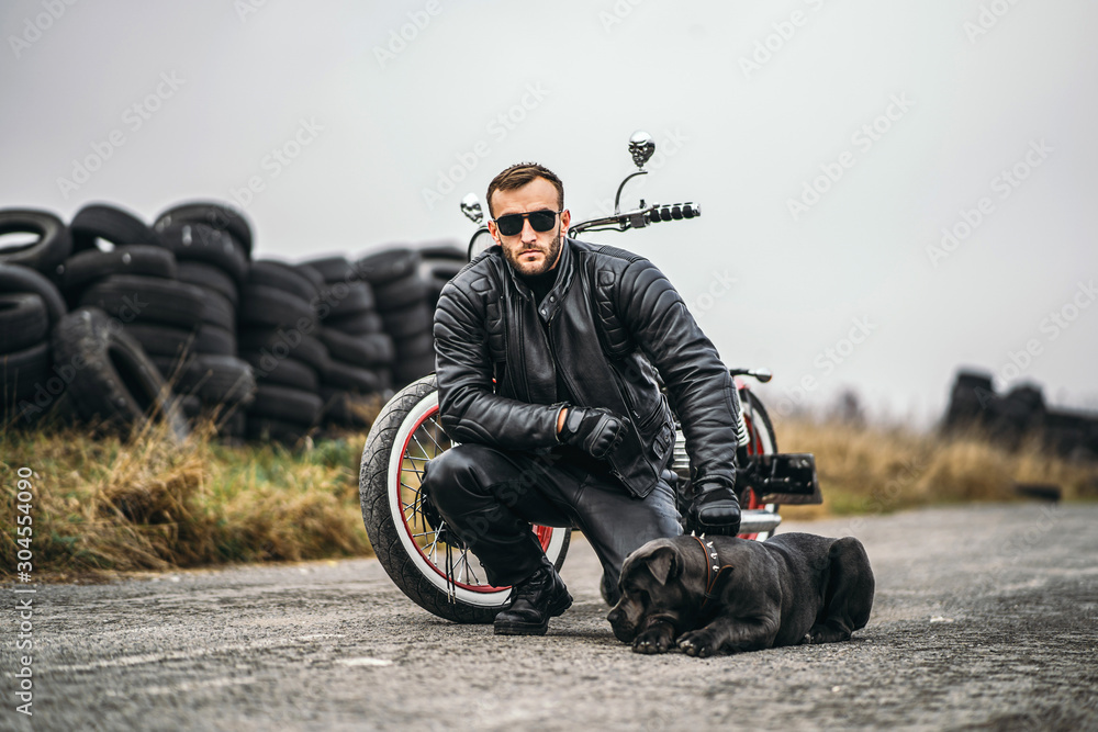 Obraz premium Biker in a leather suit crouched near his dog and red motorcycle on the road. Many tires on the background