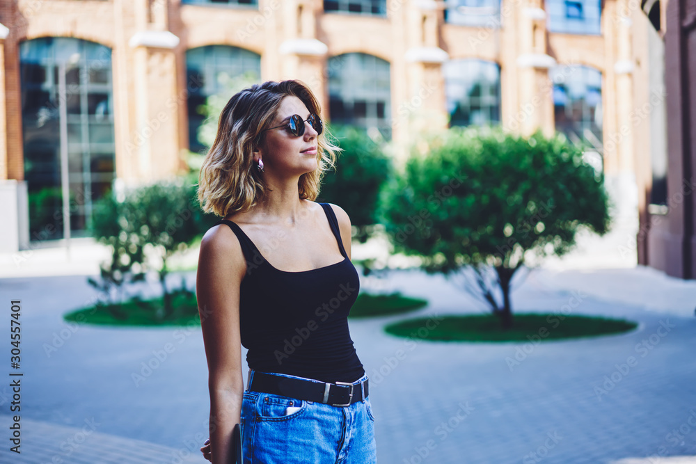 Positive female traveller in fashionable sunglasses enjoying journey during summer holidays, attractive stylish woman spending free time in college campus, concept of recreating and vacations