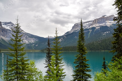 View across the beautiful Emerald Lake through pine trees towards the icy rocky mountains in the distance on a cloudy day.
