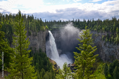 Helmcken Waterfall towers 141 meters above Murtle River with a constant flow of water gushing over the rocky cliff face feeding the dense pine forest it sits within on a partially sunny day in Canada.