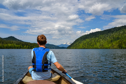 A young man dressed in blue with a life jacket on paddles his canoe along the calm Clearwater Lake towards the distant snow capped mountains on a partially cloudy day. © Hal Photography