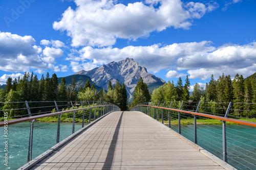 A wooden pedestrian bridge with wire tension cables and handrails on both sides with a view of a dense pine forest the rocky Cascade Mountain in the background on this partially cloudy day.