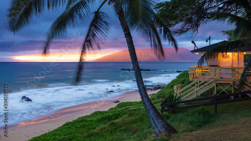 Fototapeta Naklejka Na Ścianę i Meble -  Kamaole Beach Sunset Maui. Sunset glow on the west Maui mountains, swaying palm trees in the tropical breeze, waves crashing on the sandy beach and the lifeguard hut illuminated ready for the the nigh