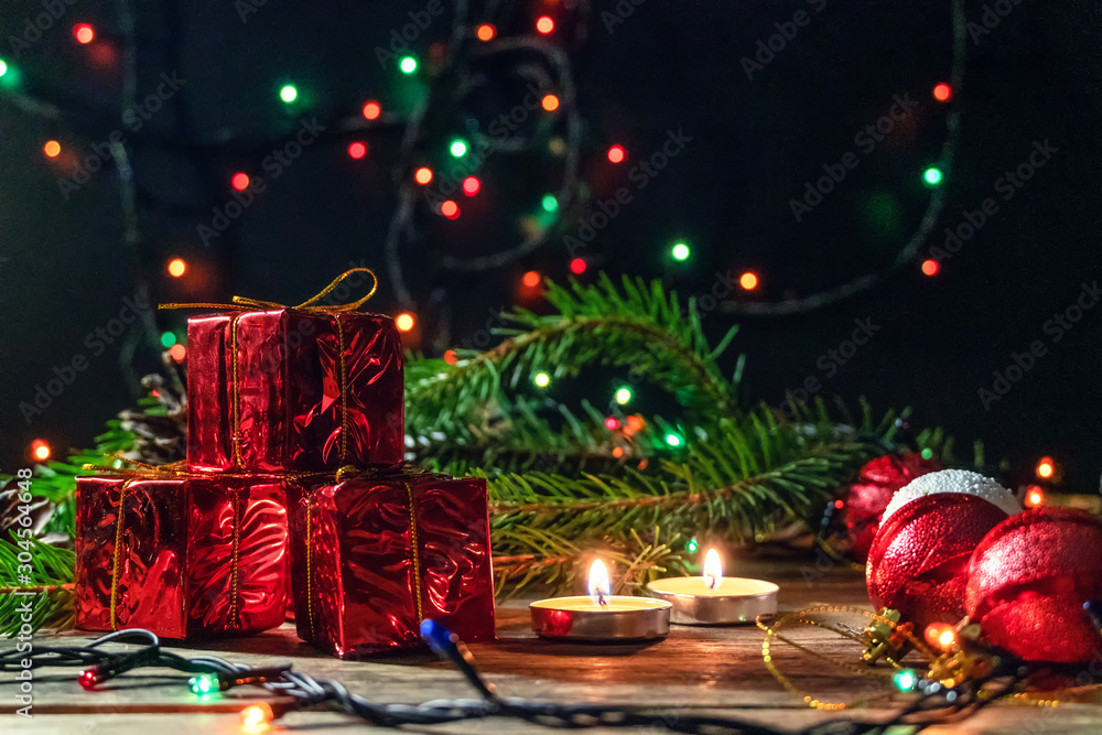 Red mini gifts on a wooden background among fir branches, lighted candles in a dark room. Beautiful, magical Christmas background with copy space.