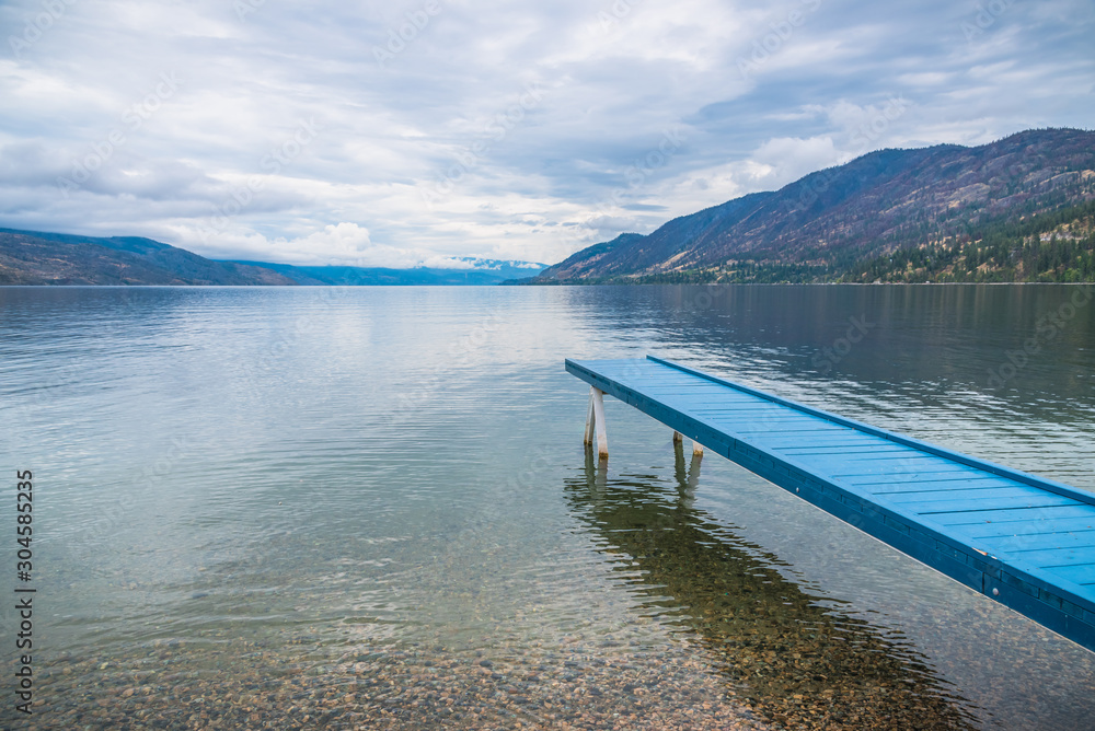 Vibrant blue painted dock extending over water toward view of mountains and overcast sky at Okanagan Lake