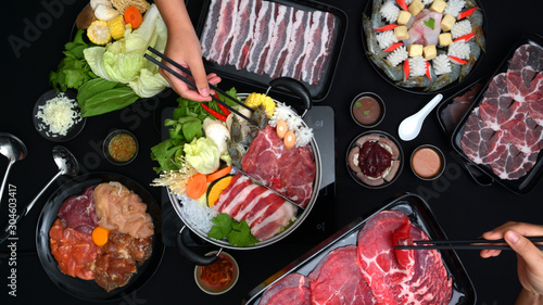 Top view of people eating Shabu-Shabu in hot pot with fresh sliced meat, sea food, and vegetables with black background
