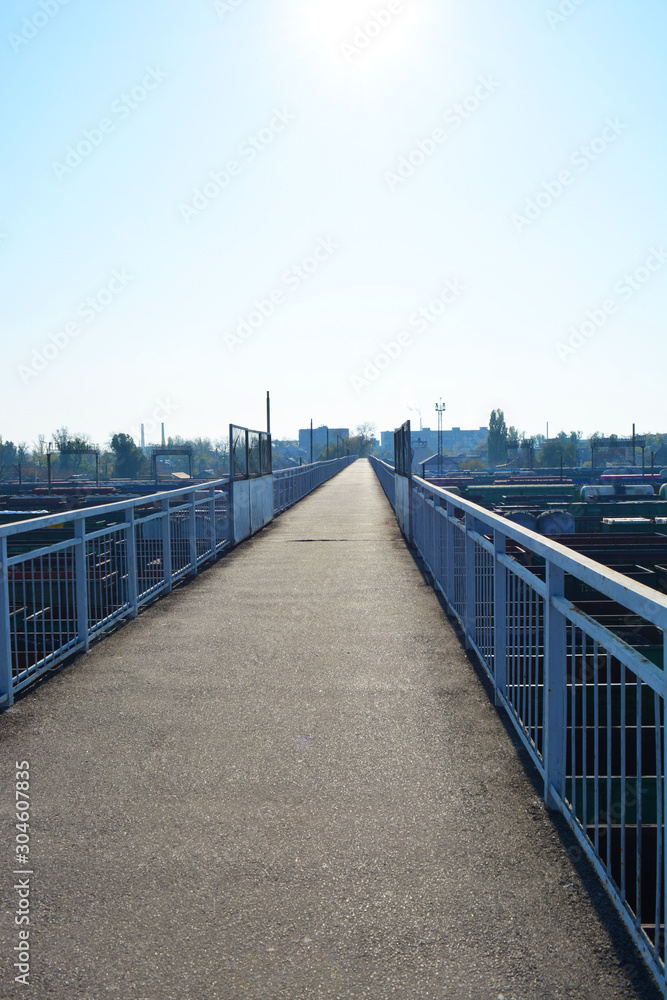 A narrow gray reinforced concrete bridge over a junction of railways and freight trains lit by the bright sun with blue sky.