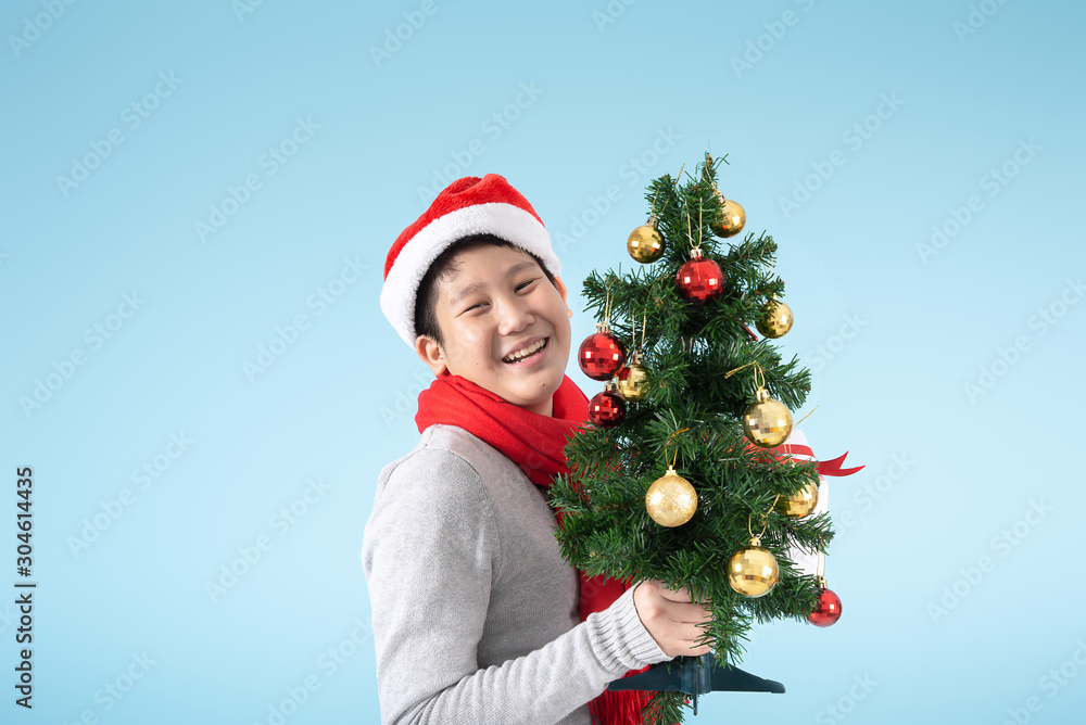 Happy Asian preteen boy holding Christmas tree and gift box on blue background, Christmas concept.