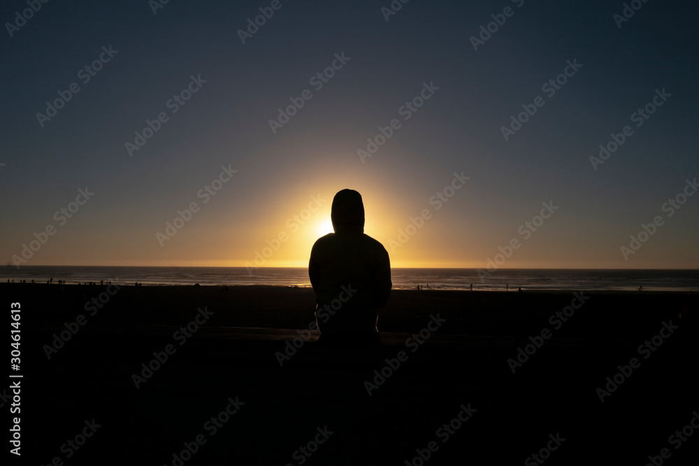 Silhouette of woman sitting at the beach by ocean in hooded jacket 