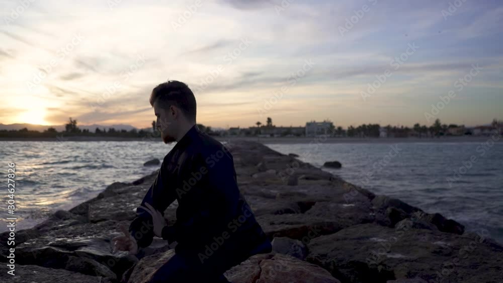 Backlight of the sun in Solow motion of a man, he meditates in tai chi style alone and and surrounded by the sea. Tai Chi training at sunset with typical tai chi dress. Backlight of the sun in Solow motion of a man, he meditates in tai chi style alone and and surrounded by the sea. Tai Chi training at sunset with typical tai chi dress.