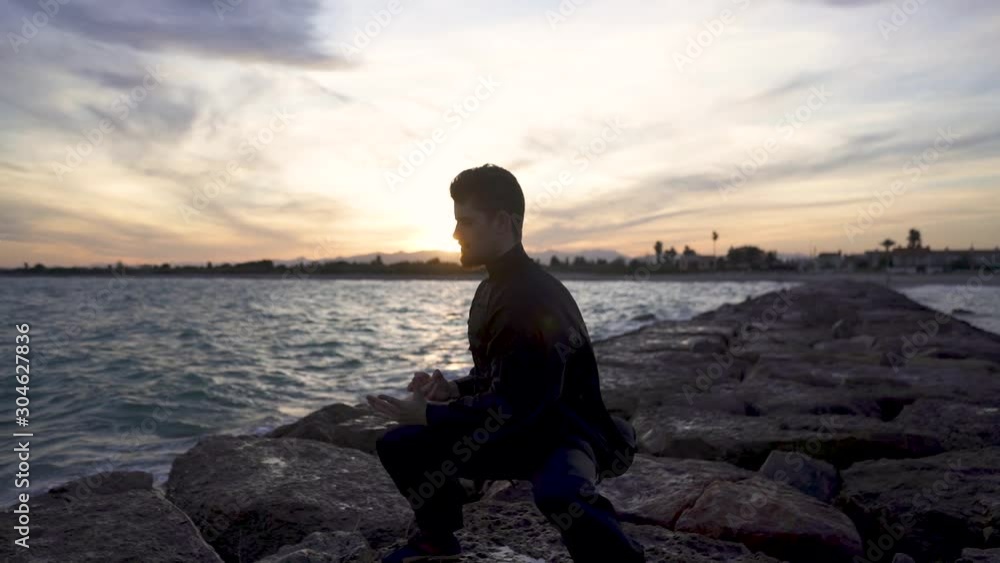 Backlight of the sun in Solow motion of a man, he meditates in tai chi style alone with the sea. Tai Chi training at sunset with typical tai chi dress. Backlight of the sun in Solow motion of a man, he meditates in tai chi style alone with the sea. Tai Chi training at sunset with typical tai chi dress.