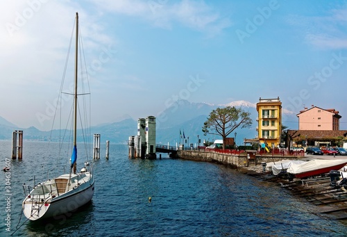 Lakeside scenery of Varenna in springtime, a beautiful village by Lake Como in Lombardy, Italy with a view of sailboats parking at the pier by lakeshore & snow capped mountains in distant background