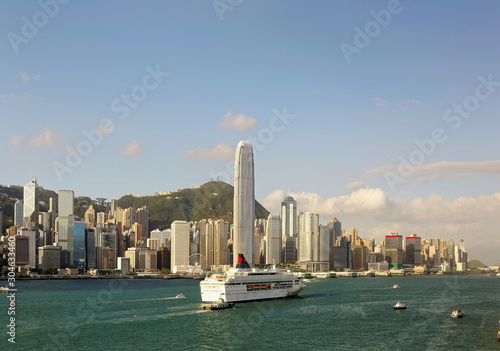 Boats & ships navigate in Victoria Harbor, Hong Kong, with the prominent landmark International Finance Center (IFC) standing among crowded skyscrapers & Victoria Peak in background under sunny sky