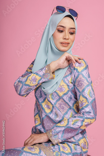 Beautiful female model wearing batik design  baju kurung  with hijab  sitting and posing on a chair isolated over pink background. Eidul fitri fashion and beauty concept.