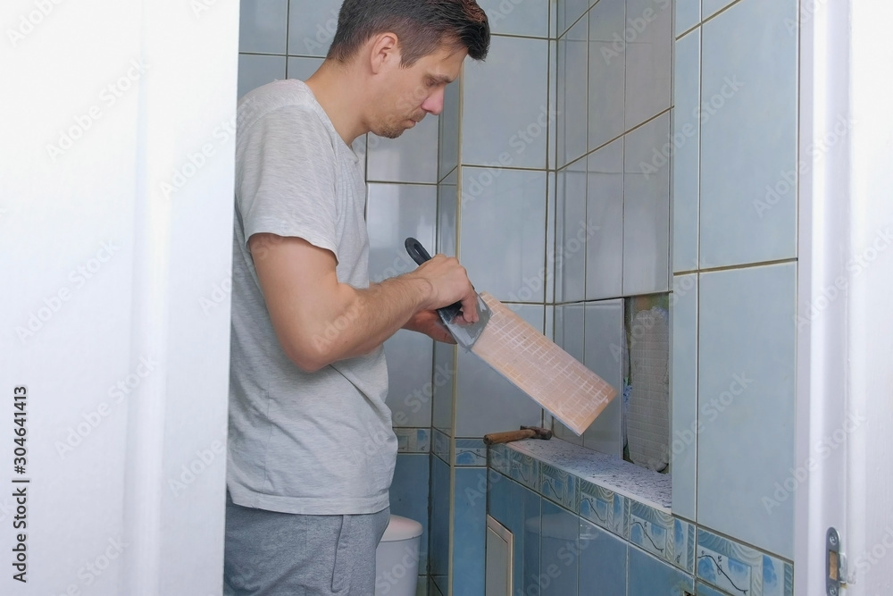 Man tiler making renovation repair at home, he removes tiles from wall in toilet using tool spatula. Construction works at home. He removes seams between tiles to remove it. Man tiler making renovation repair at home, he removes tiles from wall in toilet using tool spatula. Construction works at home. He removes seams between tiles to remove it.