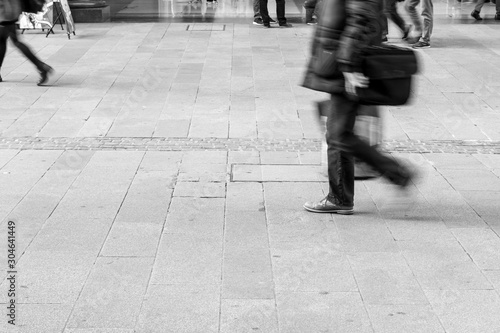 Black and white Picture of footpath as many people walk cross by. People on the street. Picture focusing on concrete with pedestrian’s shadow on. Silhouettes and Shadows of a person on the street.