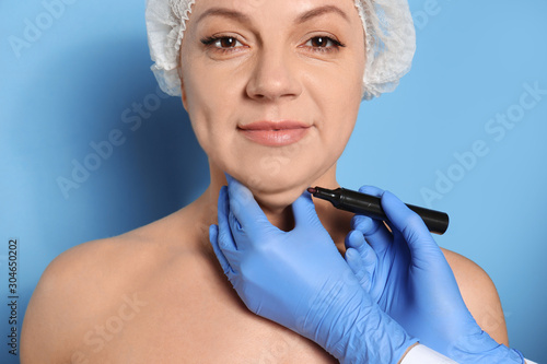 Surgeon with marker preparing woman for operation against blue background. Double chin removal