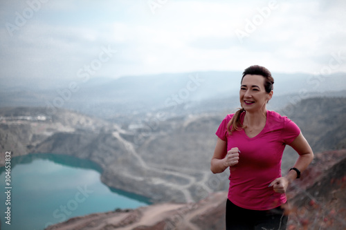 Miiddle aged woman running at the nature outdoor over mountain peaks and the lake. The traveller dressed sport wear.