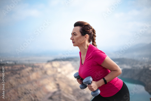 Fototapeta Naklejka Na Ścianę i Meble -  40s years woman dressed sportswear, pink topic and black fitness leggings, doing streching  sport exercise with dumbbell outdoor against the heart shaped  quarry .