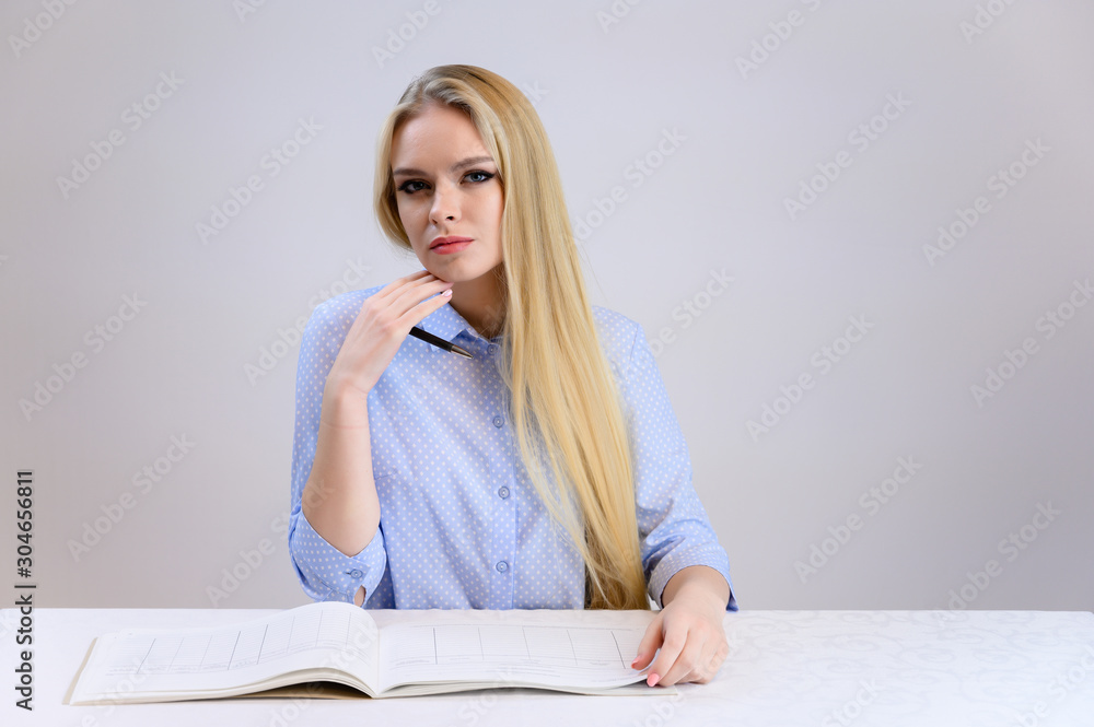 Concept cute model student secretary works sitting at a table. Close-up portrait of a beautiful blonde girl with excellent makeup with long smooth hair on a white background in a blue shirt. Concept cute model student secretary works sitting at a table. Close-up portrait of a beautiful blonde girl with excellent makeup with long smooth hair on a white background in a blue shirt.