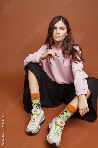 Full length shot of a dark-haired girl, sitting on a floor. She wearing pink sweatshirt, black culottes, light green artwork socks with horse jockey print  and white sneakers. 