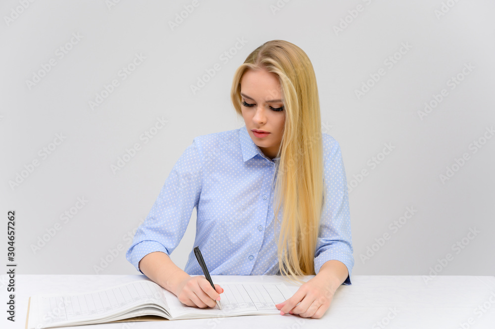 Concept cute model student secretary works sitting at a table. Close-up portrait of a beautiful blonde girl with excellent makeup with long smooth hair on a white background in a blue shirt. Concept cute model student secretary works sitting at a table. Close-up portrait of a beautiful blonde girl with excellent makeup with long smooth hair on a white background in a blue shirt.